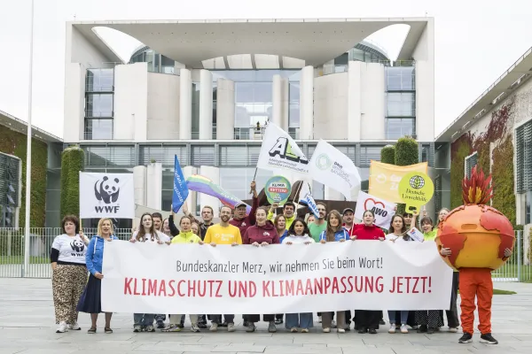 zwei Dutzend Umweltaktive vor dem Bundeskanzleramt in Berlin mit einem Banner, Aufschrift "Bundeskanzler Merz, wir nehmen Sie beim Wort! KLIMASCHUTZ UND KLIMAANPASSUNG JETZT!"; rechts im Bild, eine als brennende Erde verkleidete Person